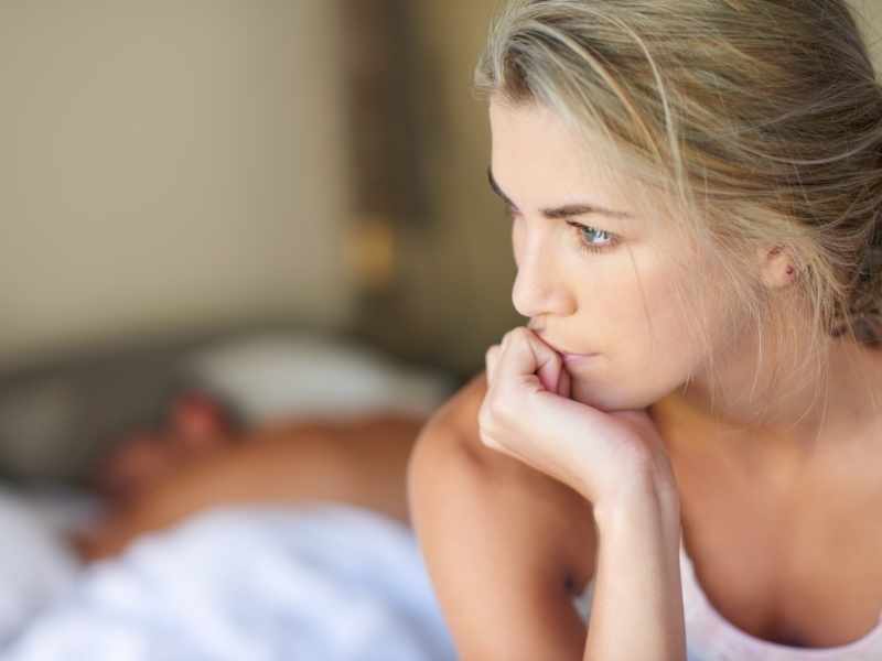 mujer sentada en la cama con su pareja al fondo, está confundida por el resultado de su prueba de embarazo casera